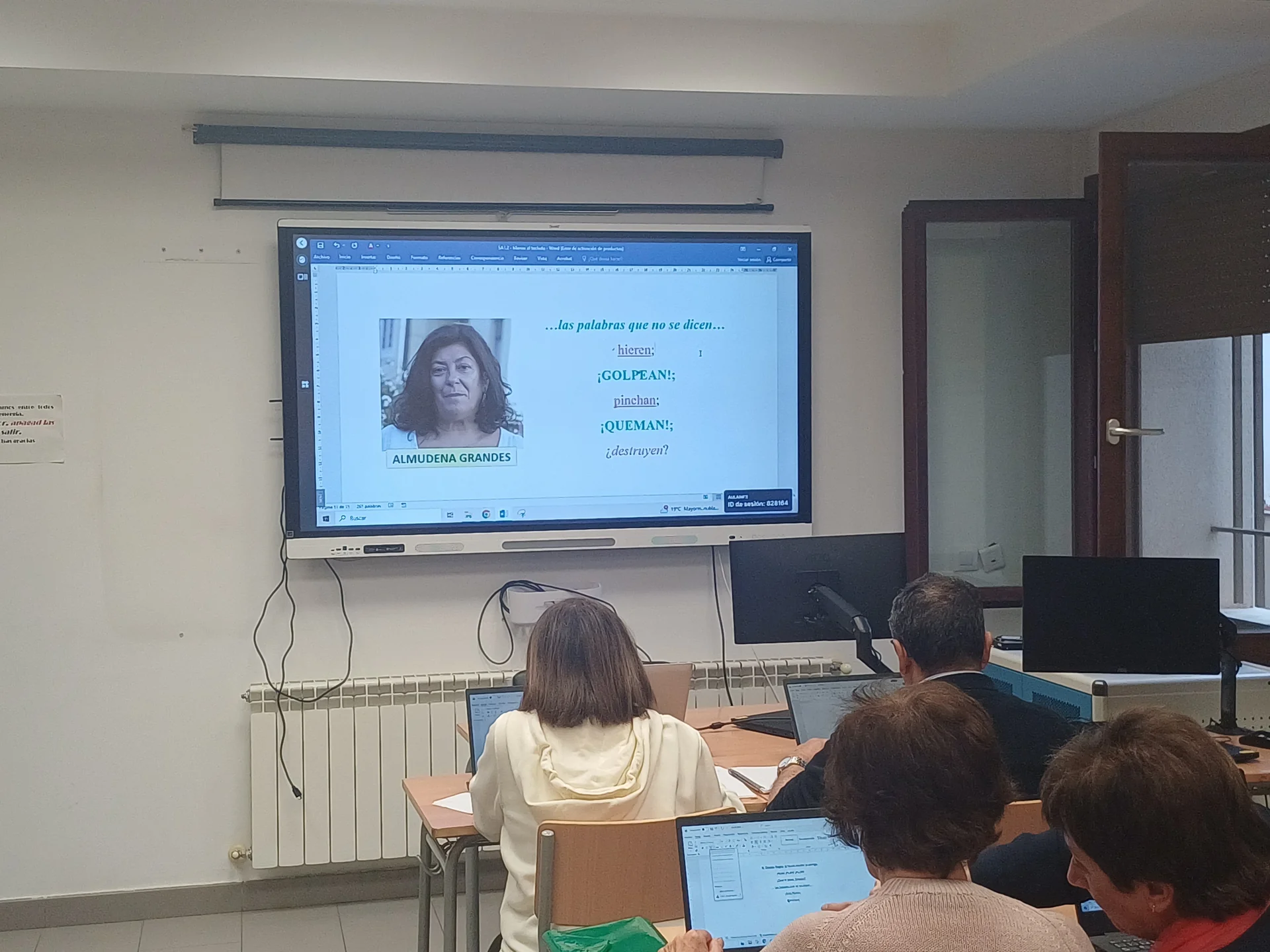 Pantalla del aula con un texto presentado junto al nombre y retrato de Almudena Grandes en Microsoft Word, durante la práctica con ordenadores.