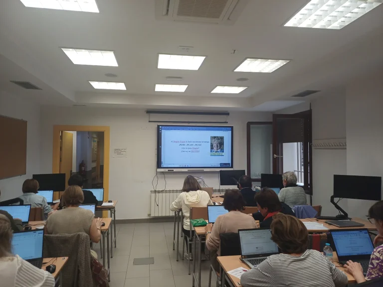 Alumnado de Competencia Digital trabajando con ordenadores portátiles mientras se muestra un texto de Gloria Fuertes en la pantalla del aula.